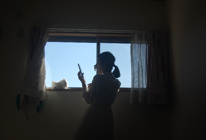 窓辺でスマホを操作している女性の後ろ姿、友達の婚約者が自分の連絡先を聞いてきた理由を知りたいと思っている。